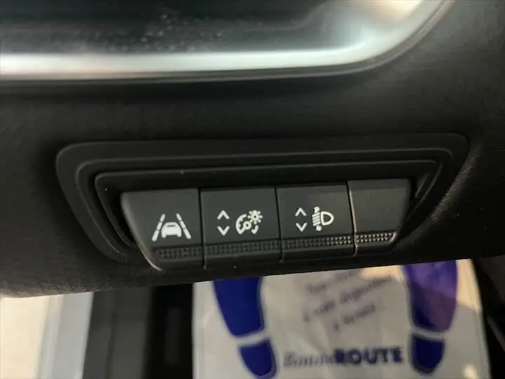 Boutons de réglage des aides à la conduite et des phares sur la planche de bord d'une Renault Clio V blanche nacrée.