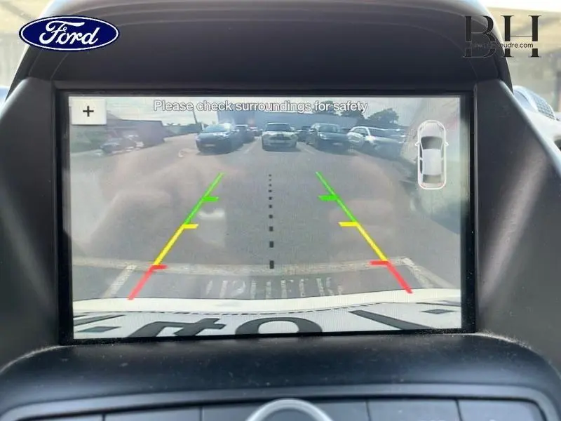 Écran de caméra de recul du Ford Kuga blanc glacier montrant l'arrière avec lignes de guidage colorées et parking visible.