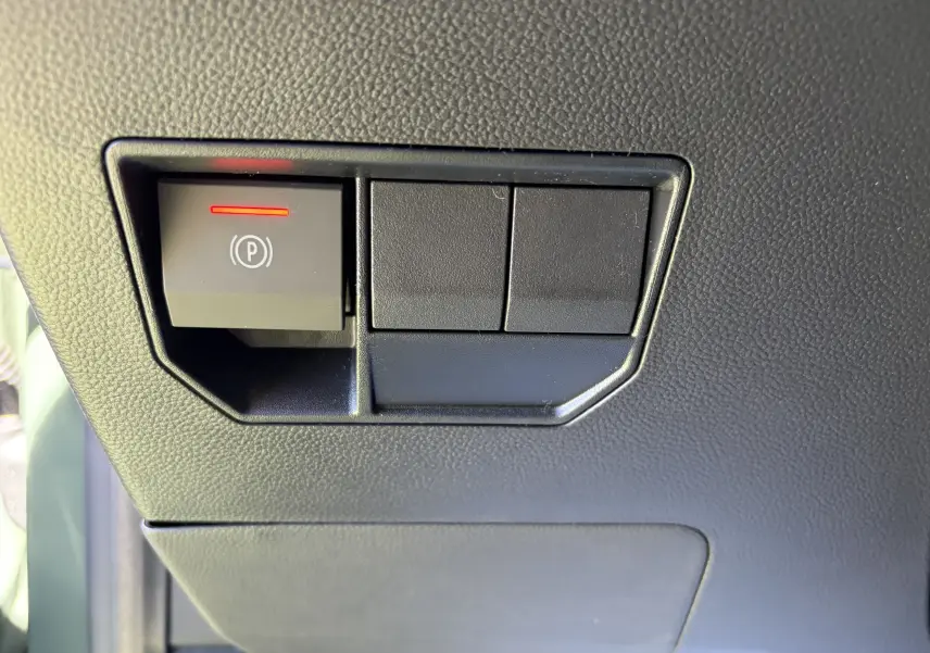 Gros plan sur le bouton de frein de stationnement électrique avec témoin rouge, tableau de bord noir du Dacia Duster III.