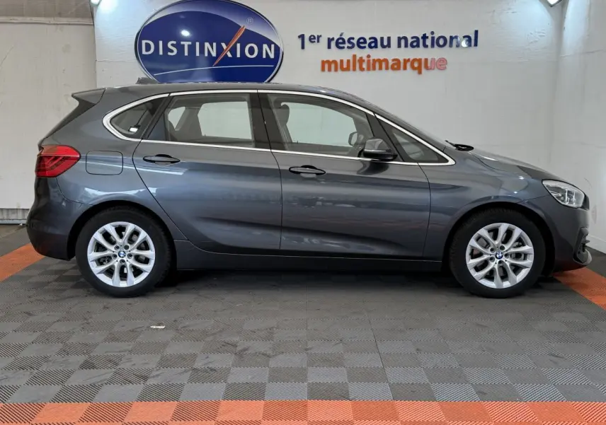 BMW Série 2 Active Tourer gris foncé vue de profil côté gauche, avec jantes alliage et logo Distinxion en arrière-plan.