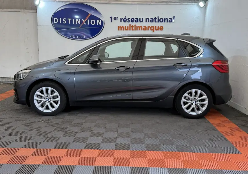 Profil côté gauche d'une BMW Série 2 Active Tourer 225xe gris foncé dans un showroom avec logo Distinxion en arrière-plan.