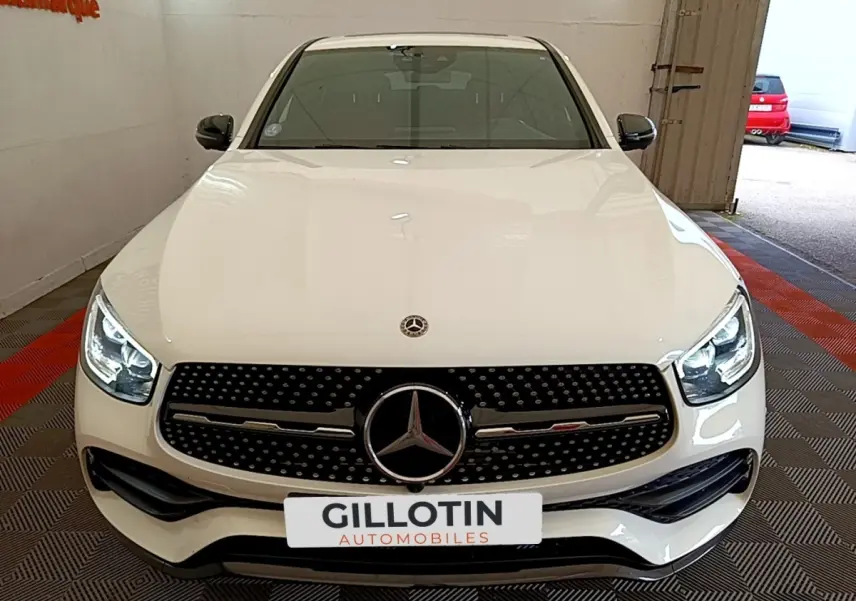 Vue avant d'un Mercedes GLC Coupé blanc avec calandre AMG noire et phares LED allumés dans un garage.