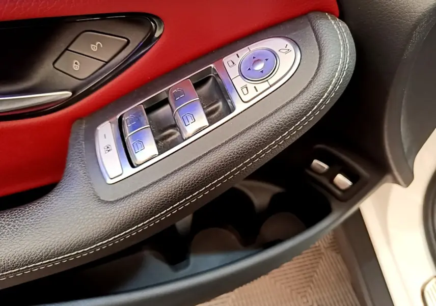 Gros plan sur la commande des vitres et rétroviseurs côté conducteur, garniture cuir noir et panneau rouge, porte blanche visible.