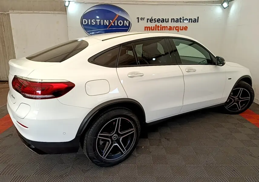 Vue 3/4 arrière droite d'un Mercedes GLC Coupé blanc avec jantes AMG noires et vitres teintées en intérieur showroom.