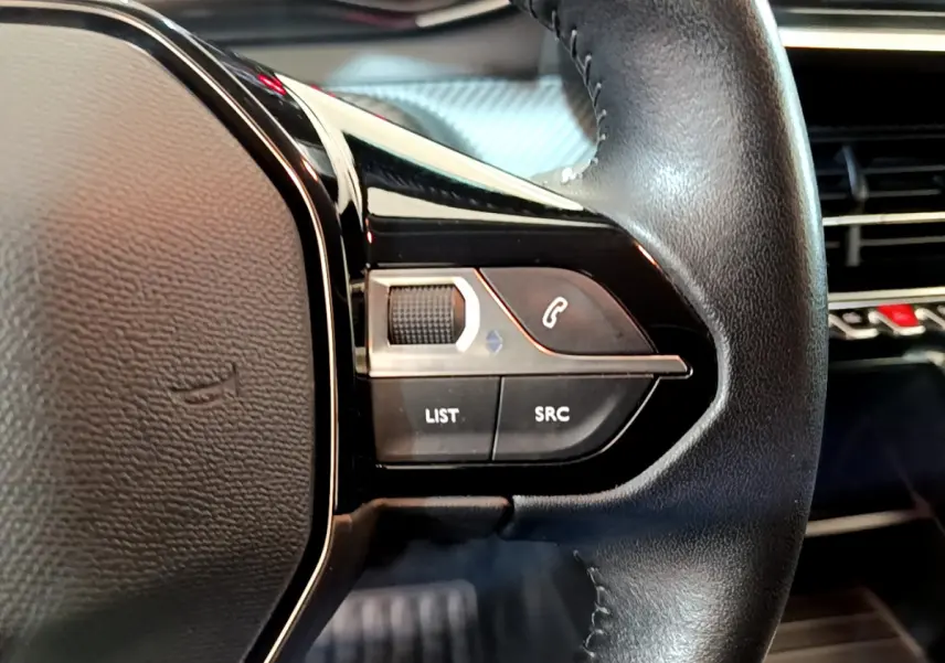 Gros plan sur les commandes gauche du volant cuir noir du Peugeot 2008 gris clair avec tableau de bord en arrière-plan.