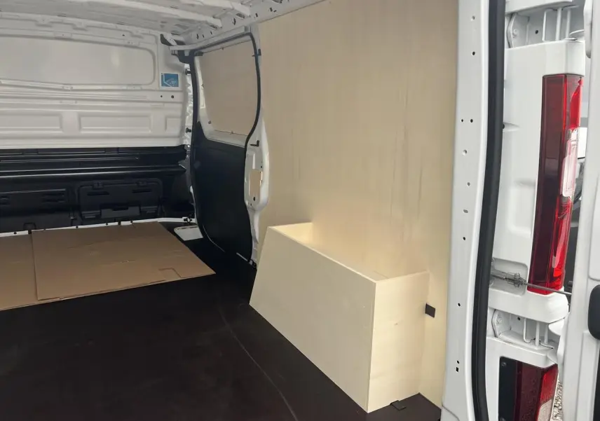 Vue intérieure arrière droite du Nissan Primastar blanc 2025 avec aménagement bois Pack Cargo et plancher noir.