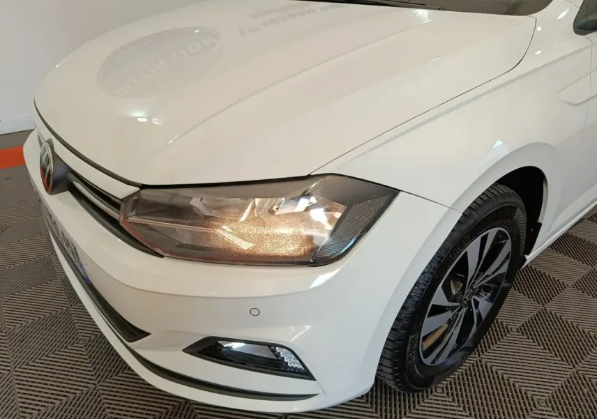 Vue 3/4 avant droit d'une Volkswagen Polo blanche avec phares allumés et jantes noires à motifs modernes.