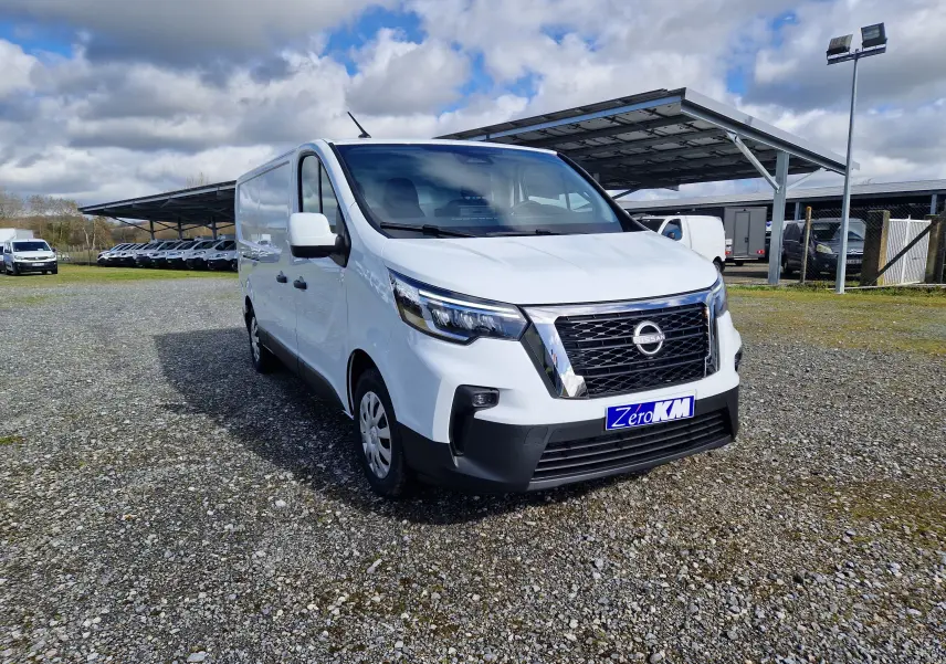 Nissan Primastar blanc en vue 3/4 avant droit sur parking extérieur avec abri métallique en arrière-plan.