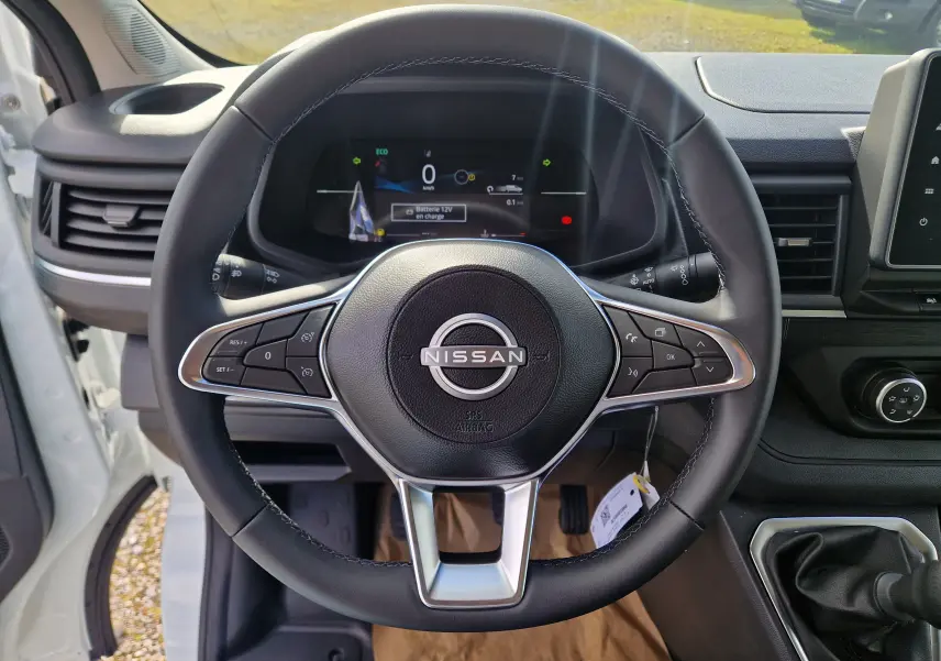 Vue intérieure centrée sur le volant noir du Nissan Primastar blanc, avec tableau de bord digital et levier de vitesses manuel visible.