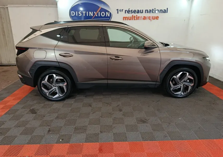 Vue latérale côté gauche d'un Hyundai Tucson marron 2022 hybride rechargeable sur sol damier gris et rouge en intérieur showroom.