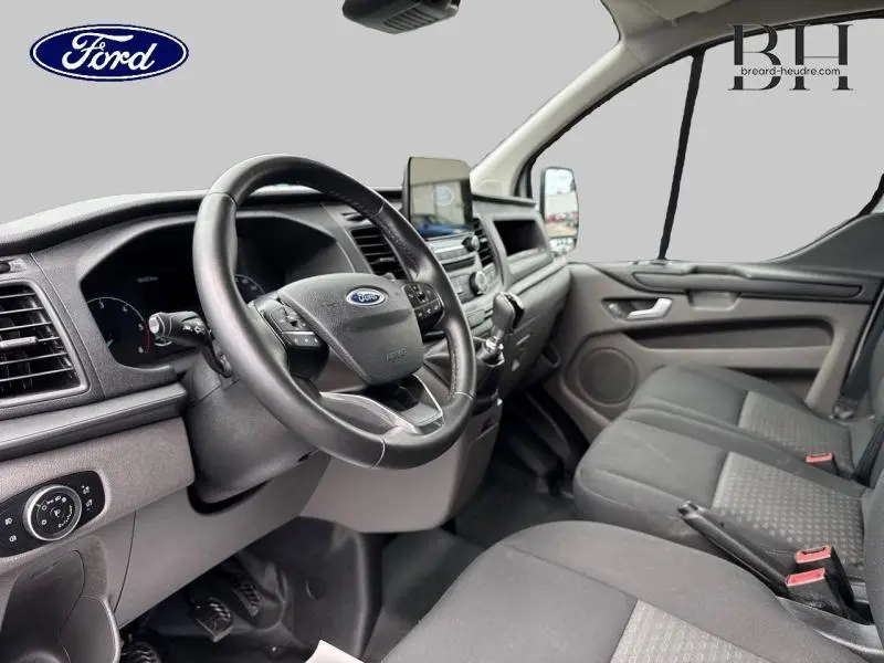 Vue intérieure côté conducteur du Ford Transit Custom blanc Glacier, montrant le volant cuir, tableau de bord et banquette avant grise.