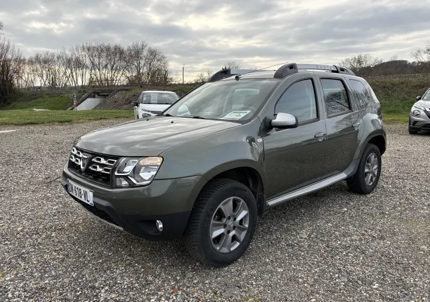 Dacia Duster 1.5 DCI 110 vert foncé en 3/4 avant droit, avec barres de toit et jantes alliage visibles.
