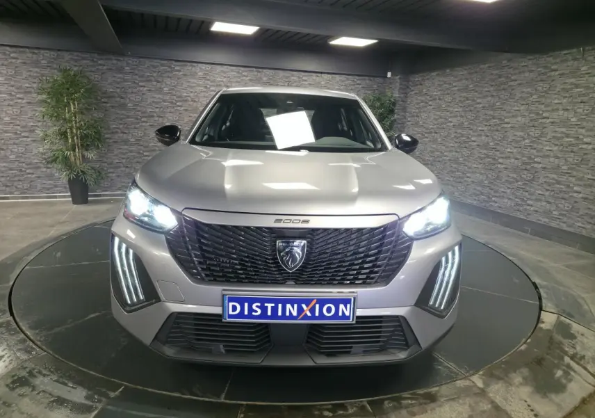 Vue de face du Peugeot 2008 gris métallisé 2025 avec calandre noire et feux LED allumés dans un showroom.