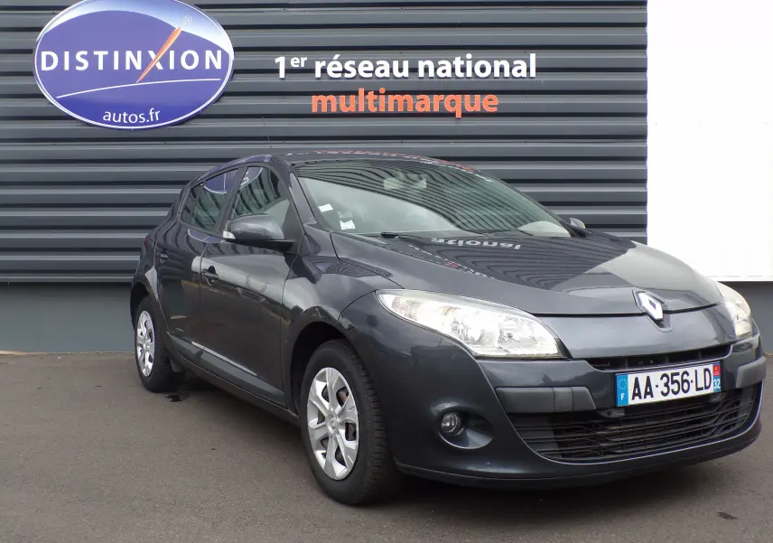 Renault Mégane III berline anthracite bleuté vue 3/4 avant droit avec phares allumés devant un mur gris.