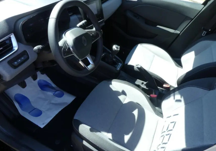 Intérieur avant droit de Renault Clio 2025, sièges gris, volant multifonction, levier de vitesse manuel et écran tactile visible.