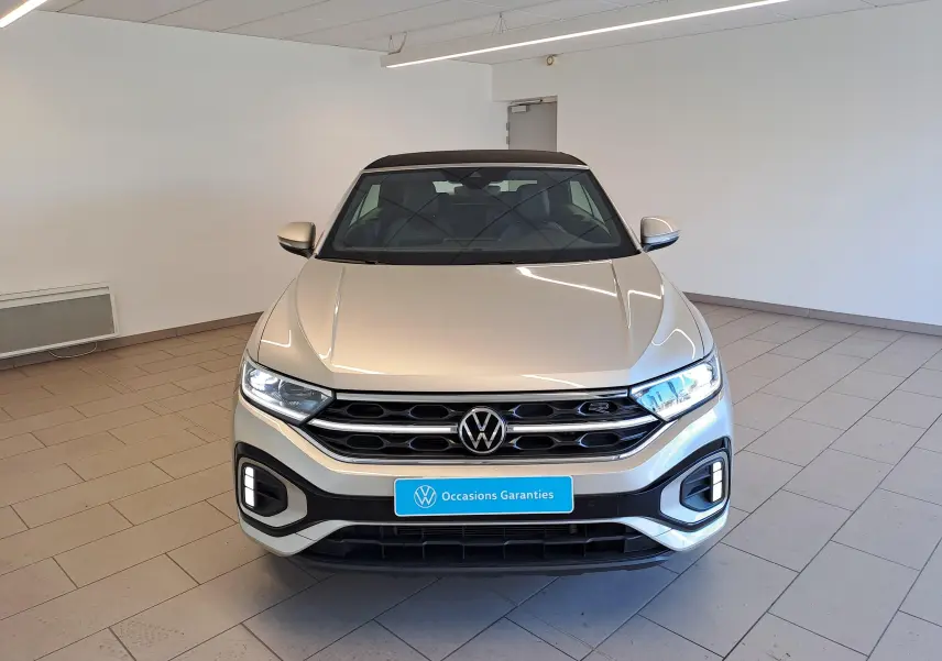 Vue frontale d'un Volkswagen T-Roc Cabriolet gris ivoire avec capote noire et calandre R-Line dans un showroom.