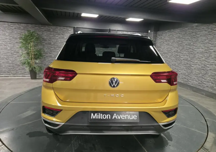 Vue arrière d'un Volkswagen T-Roc jaune Curcuma avec toit noir et feux arrière LED dans un showroom.