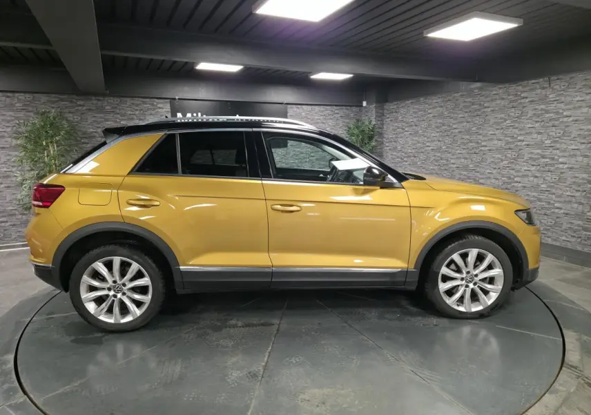Profil côté gauche d'un Volkswagen T-Roc jaune Curcuma avec toit noir et jantes alliage 18 pouces en intérieur showroom.