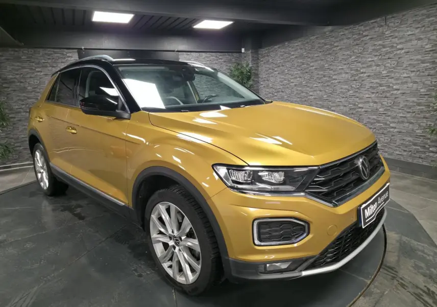 Volkswagen T-Roc jaune Curcuma en 3/4 avant droit avec toit noir et jantes alliage 18 pouces dans un showroom.