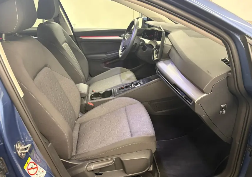 Vue intérieure côté conducteur de la Volkswagen Golf 1.5 eTSI 2025 en Bleu Anémone, sièges tissu gris et tableau de bord moderne.