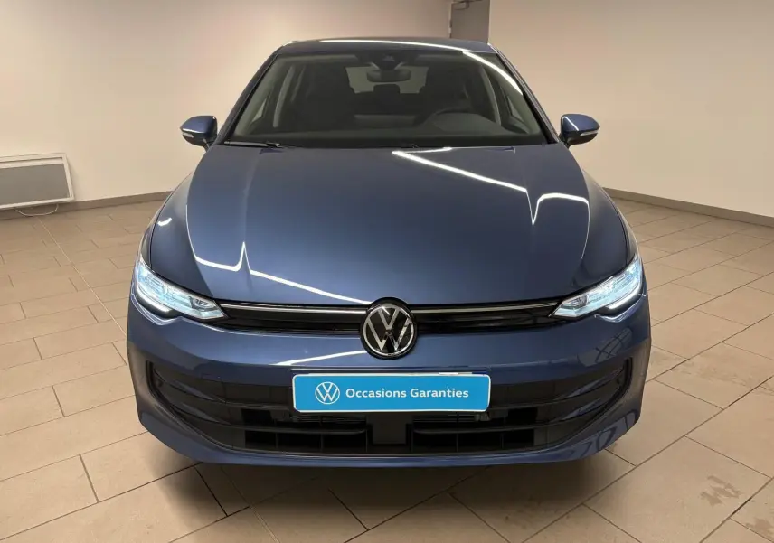 Vue frontale d'une Volkswagen Golf 1.5 eTSI 116 DSG7 VW Edition en bleu Anémone avec phares allumés dans un showroom.