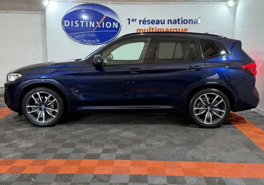 Profil côté gauche d'un BMW X3 xDrive30e bleu foncé, avec jantes alliage et toit noir en arrière-plan intérieur showroom.