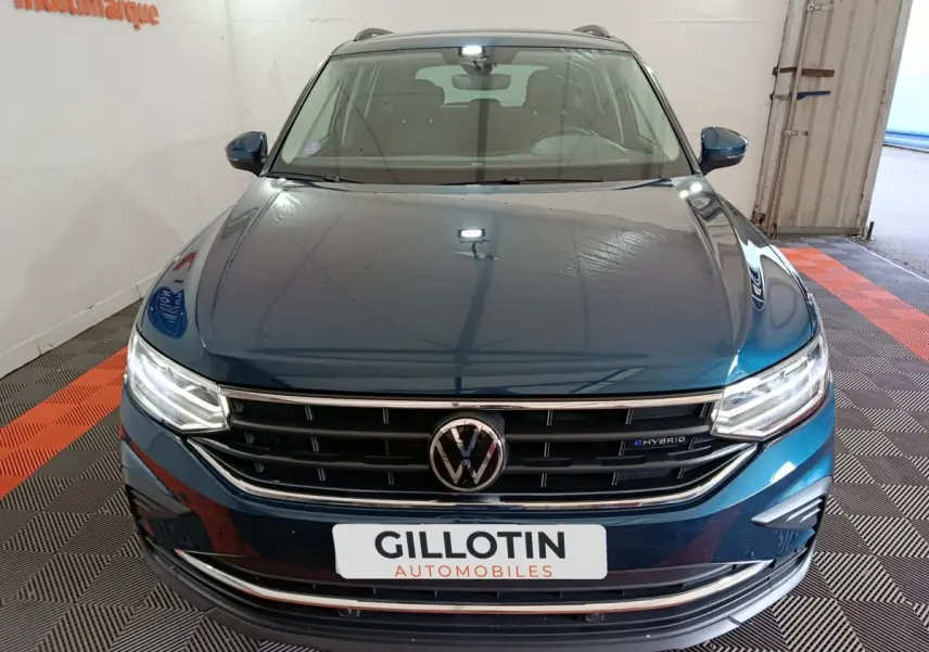 Vue avant du Volkswagen Tiguan bleu hybride avec calandre chromée et phares LED allumés dans un showroom.