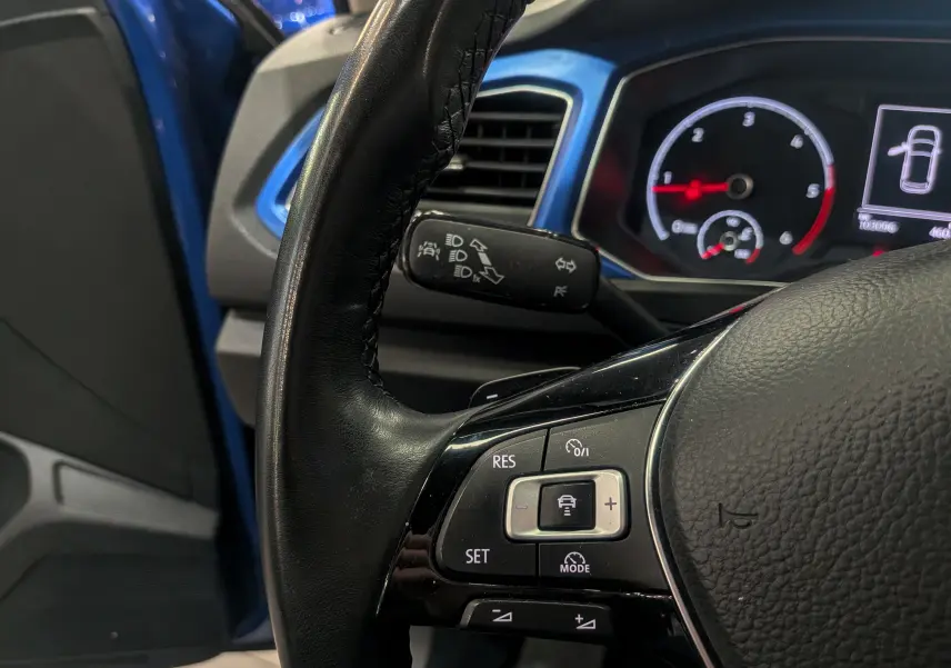 Gros plan sur le volant noir du Volkswagen T-Roc bleu Ravenna, avec commandes de régulateur et tableau de bord en arrière-plan.