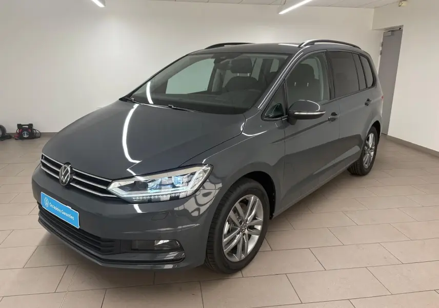 Volkswagen Touran gris dauphin en 3/4 avant droit dans un showroom, avec jantes alliage et feux LED allumés.