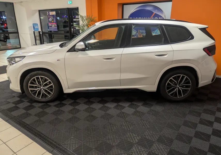Vue de profil côté gauche d’un BMW X1 blanc 2025 M Sport avec jantes alliage bicolores, garé en intérieur showroom.