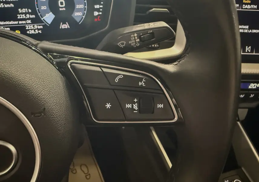 Vue rapprochée du volant noir de l'Audi A1 2024 avec commandes audio et tableau de bord numérique en arrière-plan.