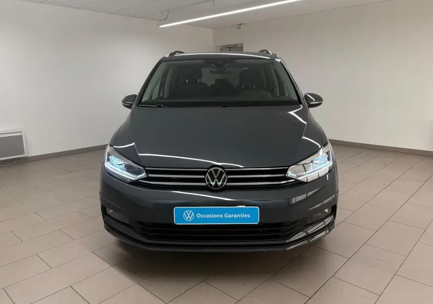 Vue frontale d'un Volkswagen Touran gris dauphin 2025 avec phares LED allumés dans un intérieur neutre.