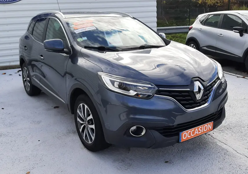 Renault Kadjar gris en 3/4 avant droit, avec calandre noire et jantes alliage bicolores visibles, stationné devant un mur blanc.