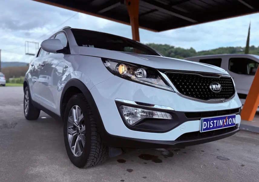 KIA Sportage blanche vue en 3/4 avant droit sous un abri avec phare avant allumé et plaque Distinxion.
