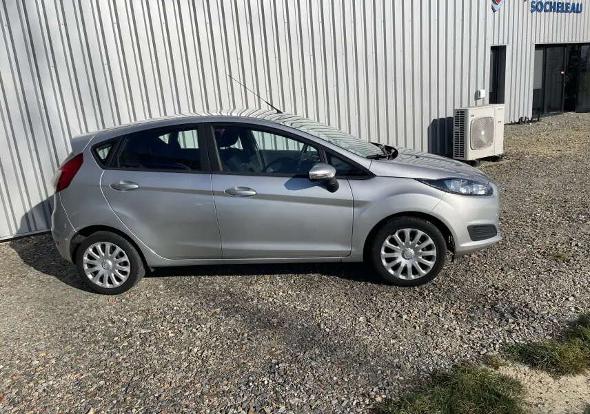 Ford Fiesta gris titane vue de profil côté gauche, garée sur un sol gravillonné devant un bâtiment industriel.
