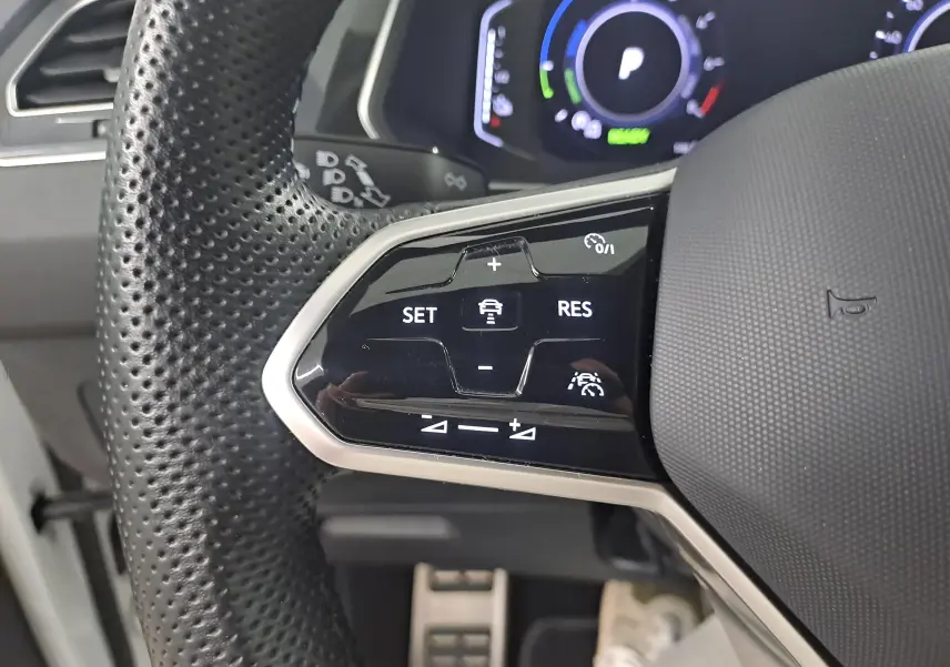 Gros plan sur les commandes du volant perforé du Volkswagen Tiguan 1.4 eHybrid blanc pur, avec affichage digital en arrière-plan.