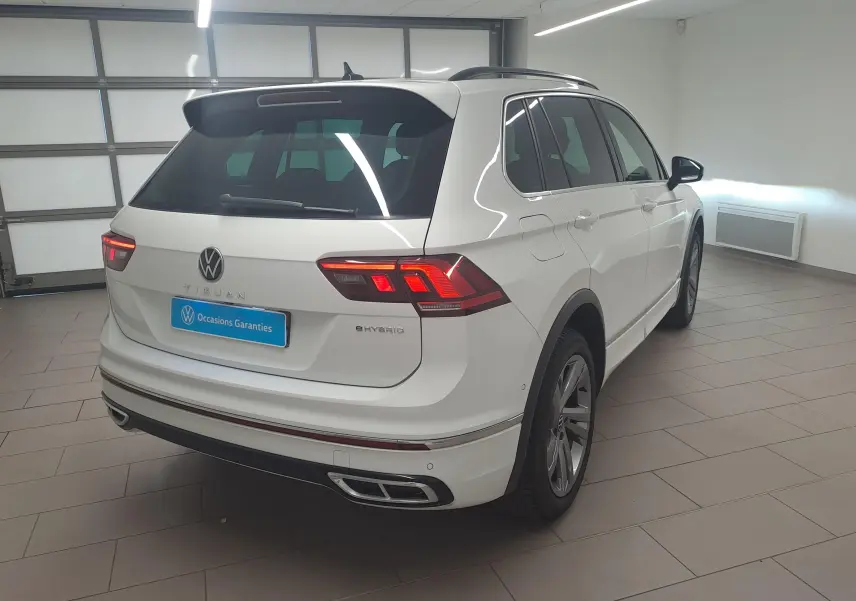 Vue 3/4 arrière droite du Volkswagen Tiguan blanc pur 2023 hybride rechargeable avec finition R-Line en intérieur showroom.