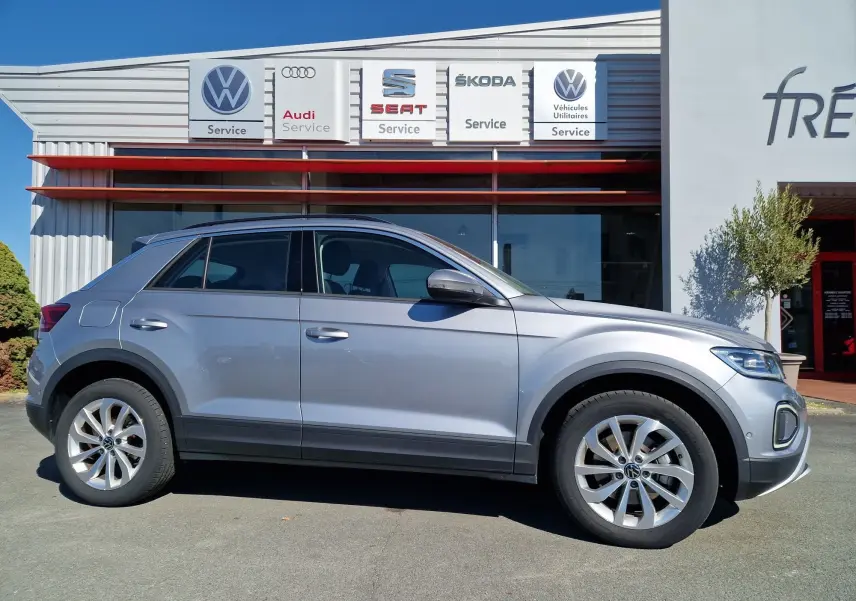 Profil côté gauche d'un Volkswagen T-Roc gris pyrite 2023 garé devant un centre de service multimarques.