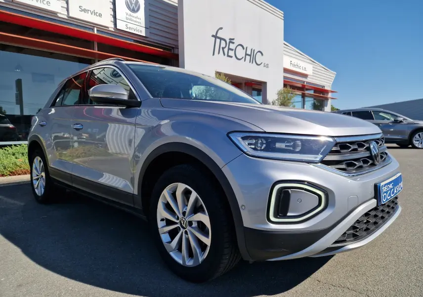 Volkswagen T-Roc gris pyrite vue 3/4 avant droit, avec phares LED et calandre distinctive, stationné devant un concessionnaire.
