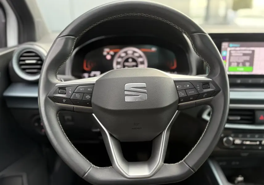 Gros plan sur le volant cuir multifonction de la SEAT Arona 2025 avec commandes intégrées et tableau de bord numérique flou.