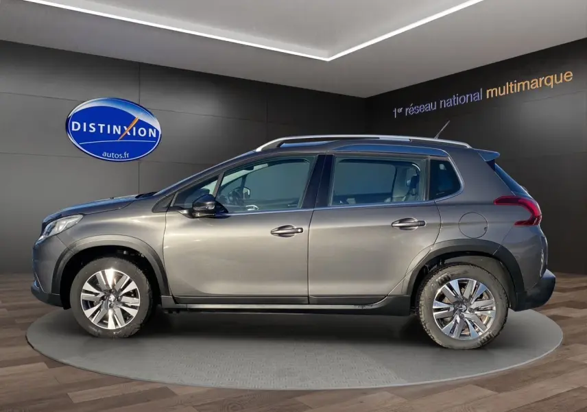 Vue de profil côté gauche d'un Peugeot 2008 gris platinium avec jantes alliage et barres de toit en intérieur showroom.