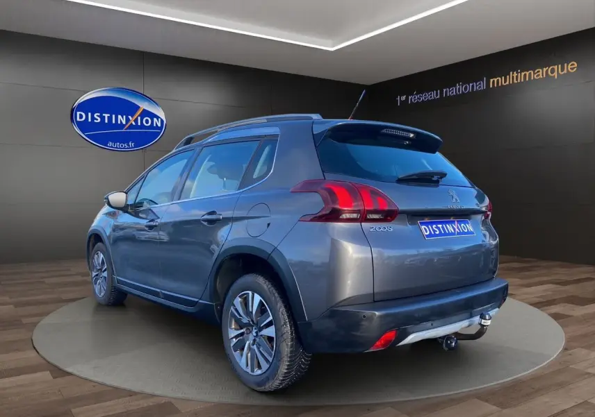 Vue 3/4 arrière droite d'un Peugeot 2008 gris platinium avec barres de toit et attelage visible en intérieur showroom.