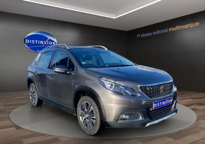 Peugeot 2008 gris platinium en 3/4 avant droit, avec jantes alliage et barres de toit visibles en showroom.