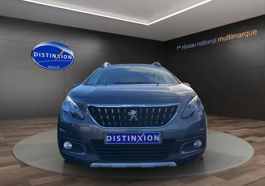 Vue frontale d'un Peugeot 2008 gris platinium avec calandre distinctive et phares allumés en showroom.