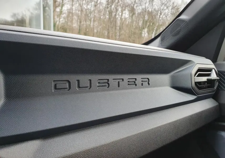 Détail intérieur du tableau de bord noir avec logo DUSTER en relief sur la partie passager du Dacia Duster 2025.
