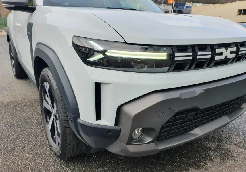 Vue 3/4 avant gauche du Dacia Duster blanc glacier 2025 avec calandre noire et phares LED allumés.