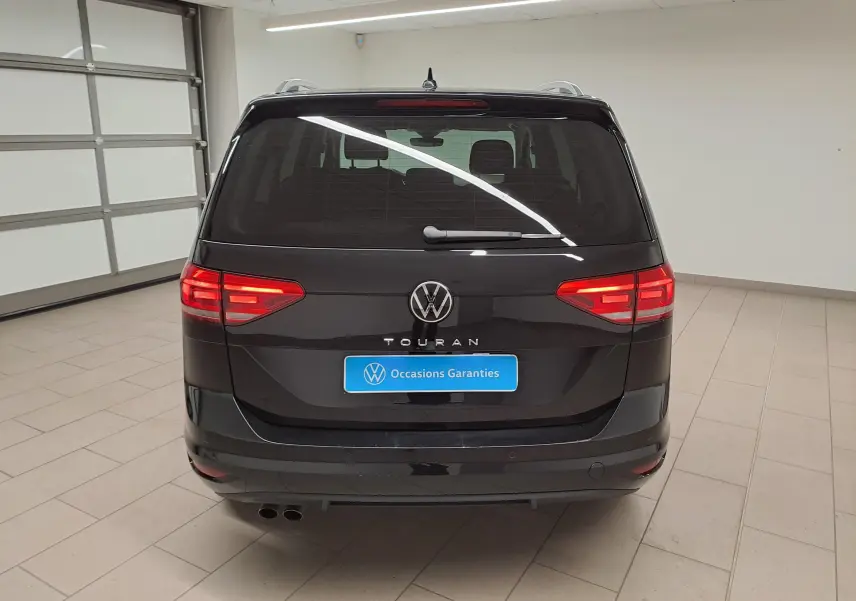 Vue arrière d'un Volkswagen Touran 2025 noir intense nacré avec feux allumés et plaque "Occasions Garantis" en intérieur.