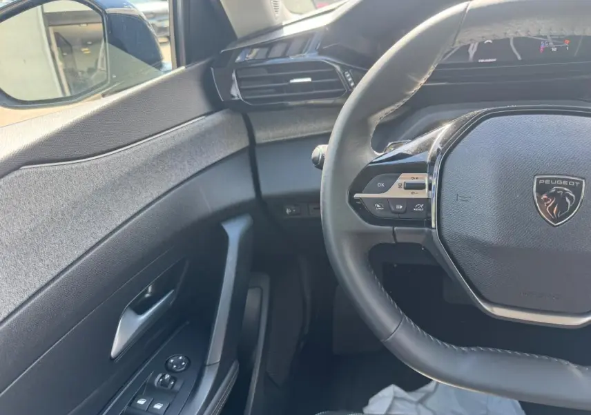 Vue intérieure côté conducteur de la Peugeot 308 Hybrid 136 Allure 2024 noire, volant multifonctions et commandes porte visibles.