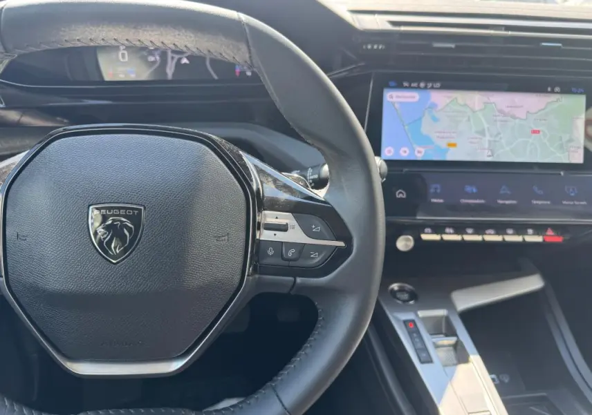 Vue intérieure centrée sur le volant Peugeot noir et le cockpit numérique avec écran tactile affichant une carte de navigation.