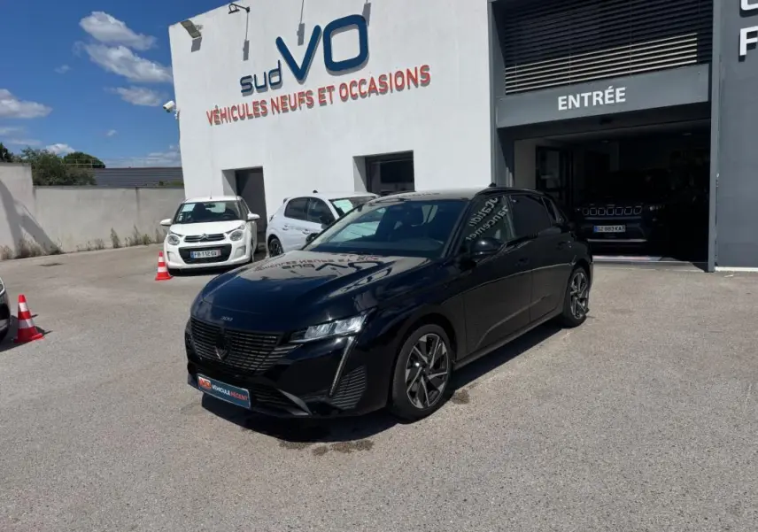 Peugeot 308 Hybrid noir vue 3/4 avant droit, jantes alliage 17 pouces, stationnée devant un concessionnaire.
